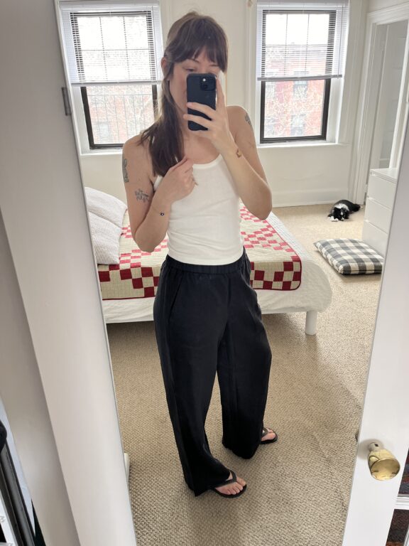 A person wearing a white tank top, black pants, and sandals takes a mirror selfie in a bedroom with a bed covered by a red and white quilt.