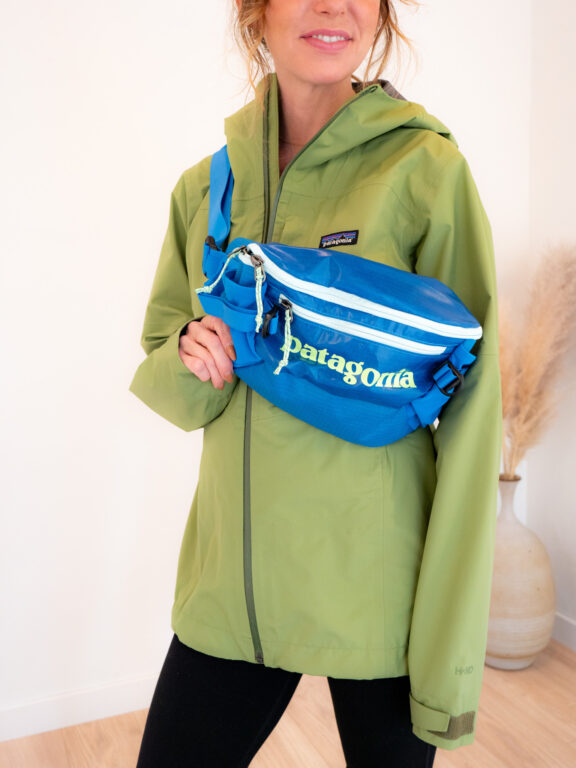 Person wearing a light green jacket and black pants, with a blue Patagonia crossbody bag, standing indoors near a beige vase with pampas grass.