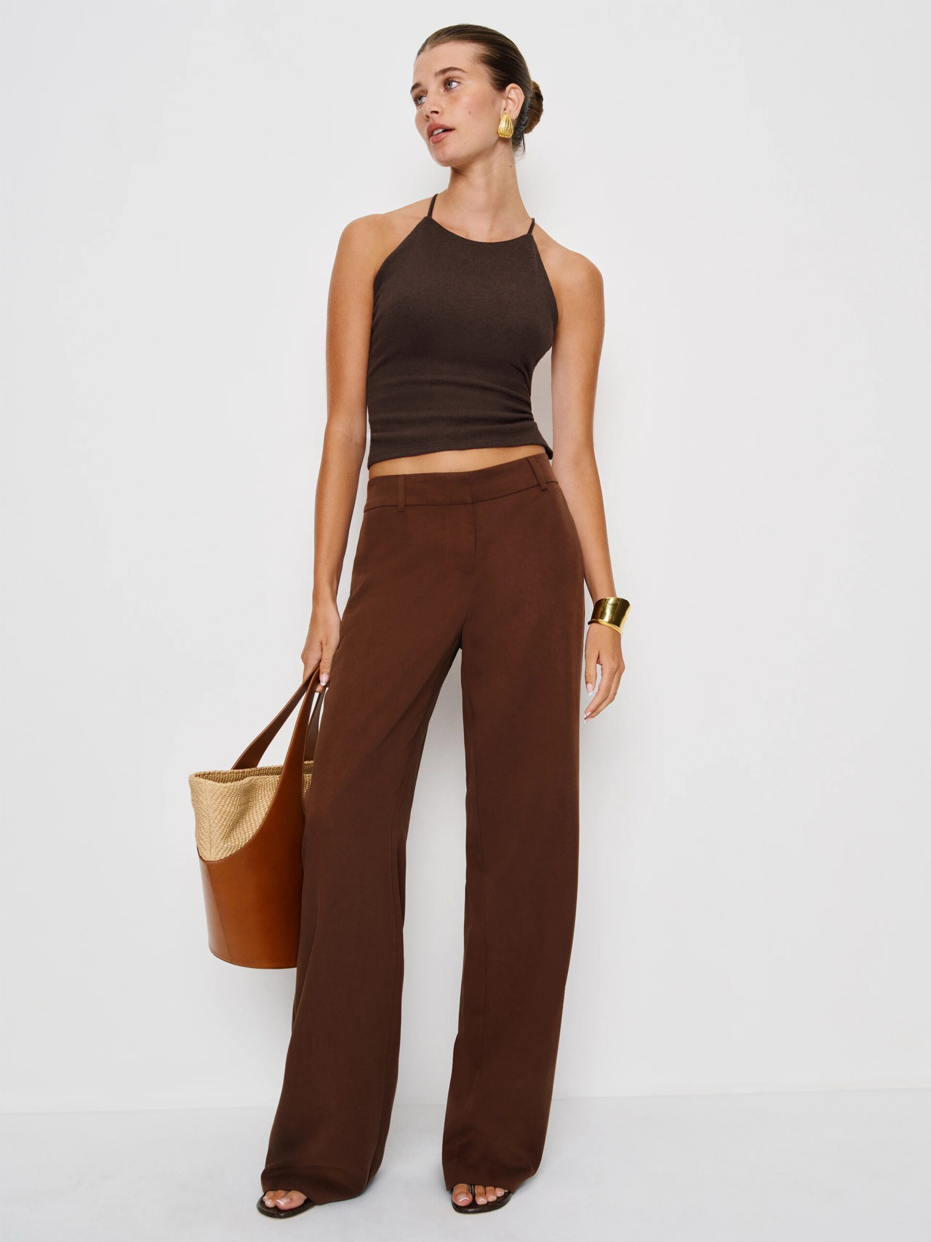 A woman stands against a white wall wearing a brown halter top, brown wide-leg pants, gold earrings, a gold bracelet, and holding a brown and straw tote bag.