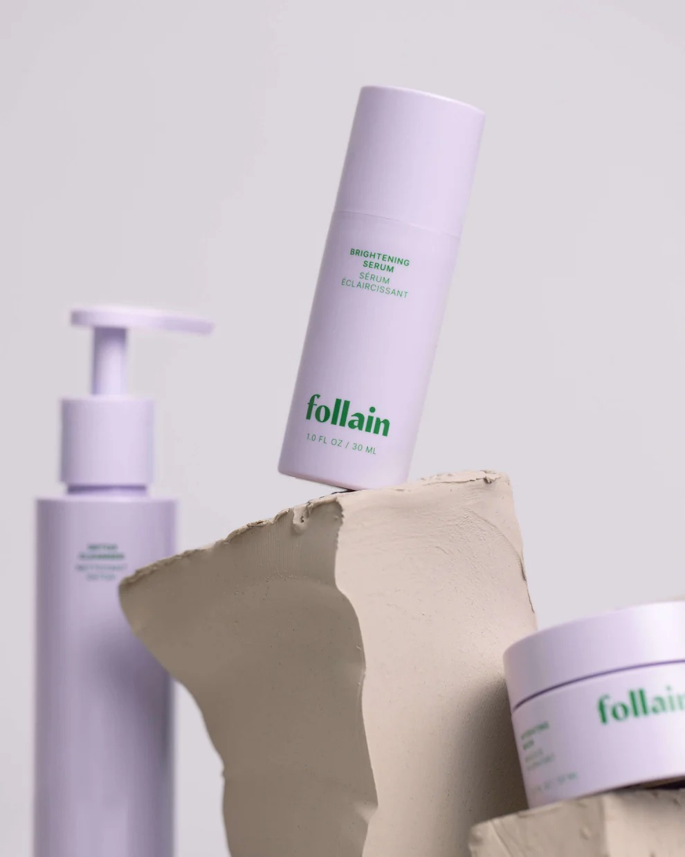 Three Follain skincare products, including a pump bottle, a serum, and a jar, are displayed on sculpted beige clay against a neutral background.