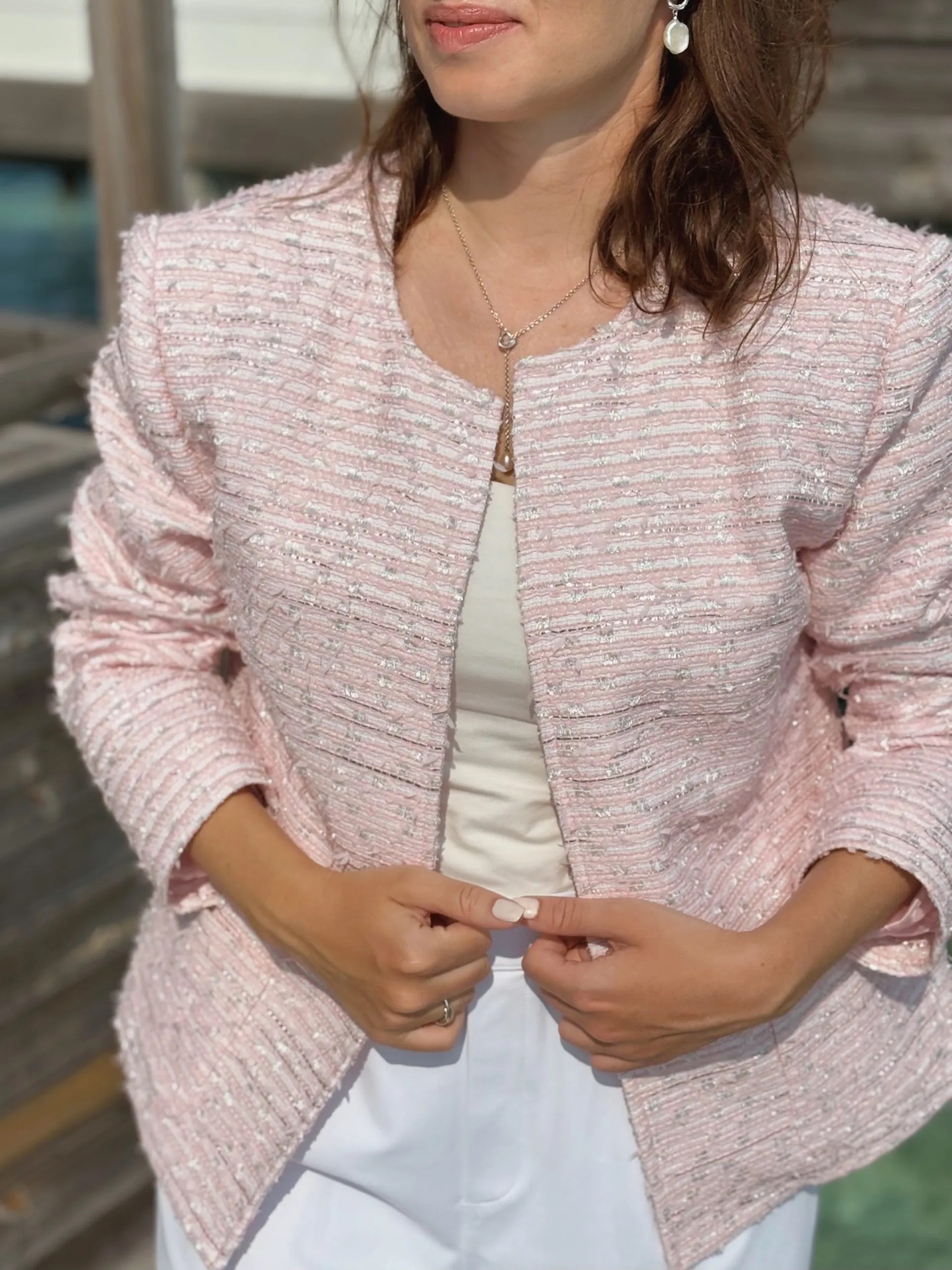 Woman wearing a light pink textured jacket over a white top and white pants, standing outdoors and holding the edges of her jacket, exuding effortless french girl fashion charm.