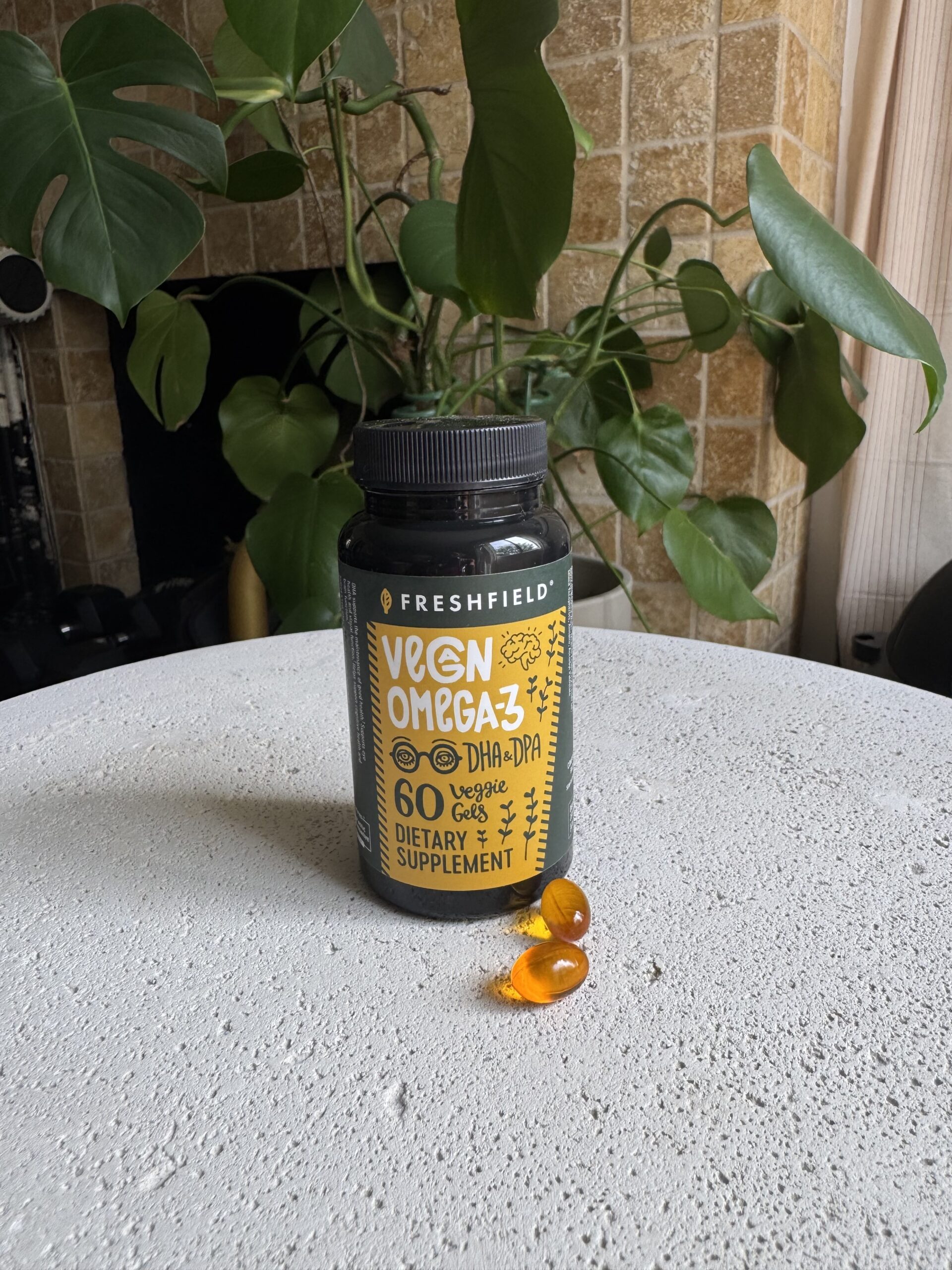 A bottle labeled "Vegan Omega-3 DHA+DPA" with 60 veggie softgels sits on a round surface beside two orange capsules; green plant in the background.