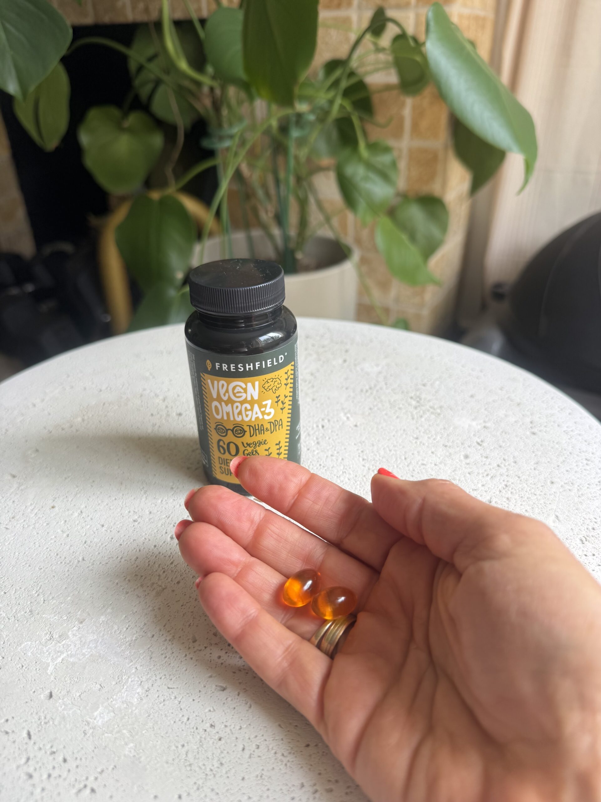 A person holds two orange gel capsules in their hand, with a bottle of Freshfield Vegan Omega-3 DHA supplement on a table in the background.