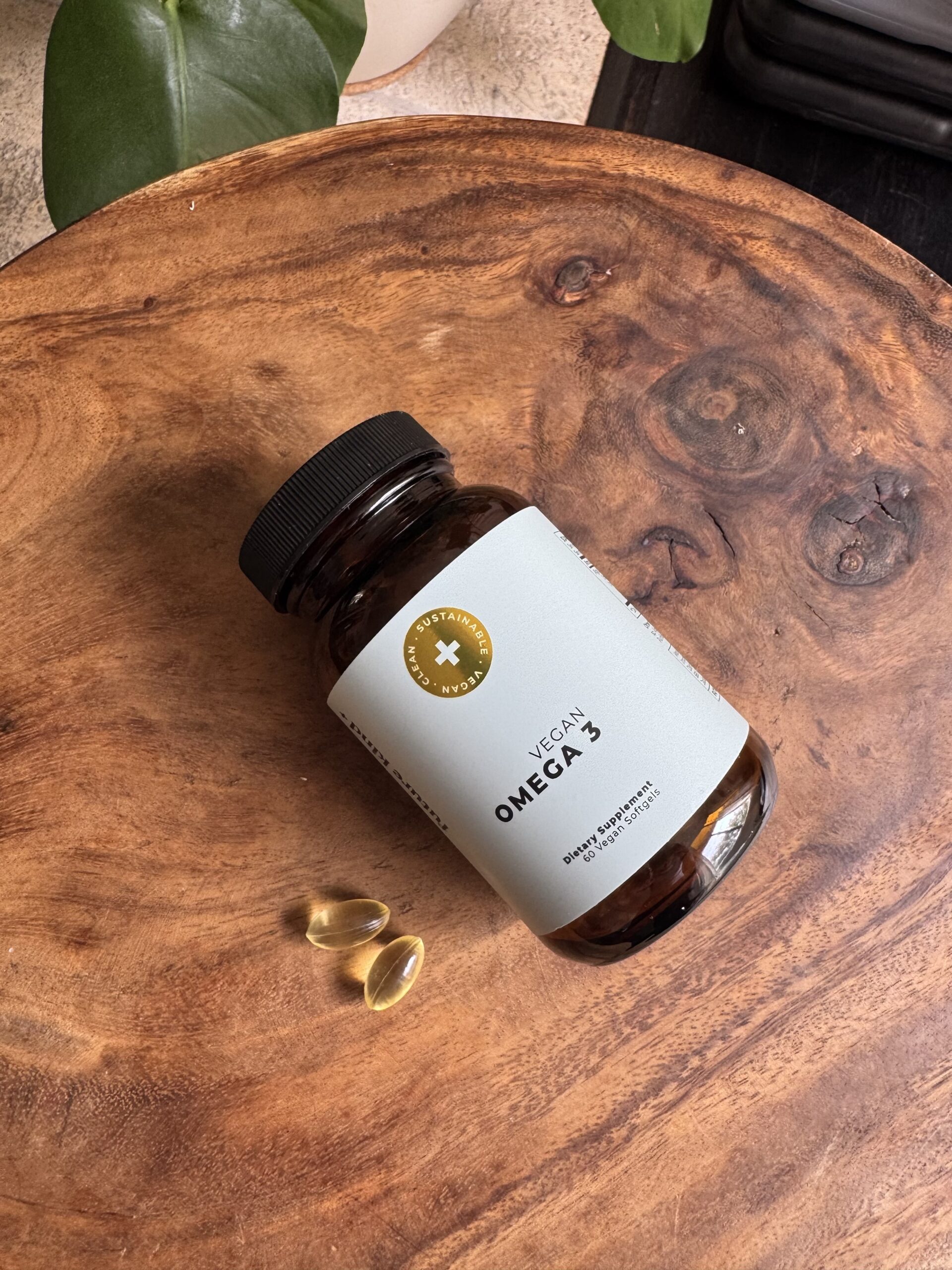 A bottle labeled "Vegan Omega 3" lies on a wooden surface with two softgel capsules next to it.