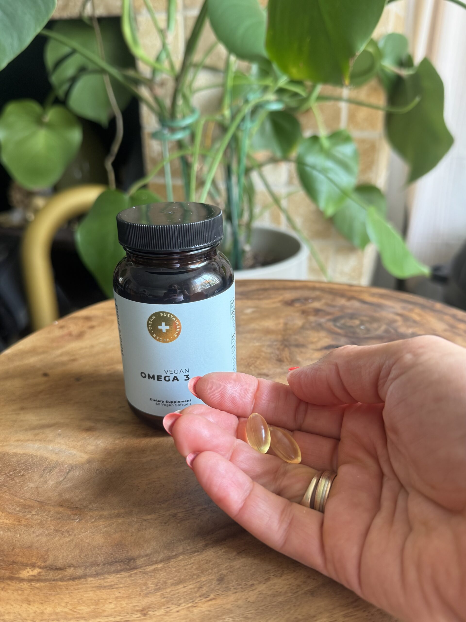 A hand holding two softgel capsules next to a bottle labeled "Vegan Omega-3" on a wooden surface, with green plants in the background.