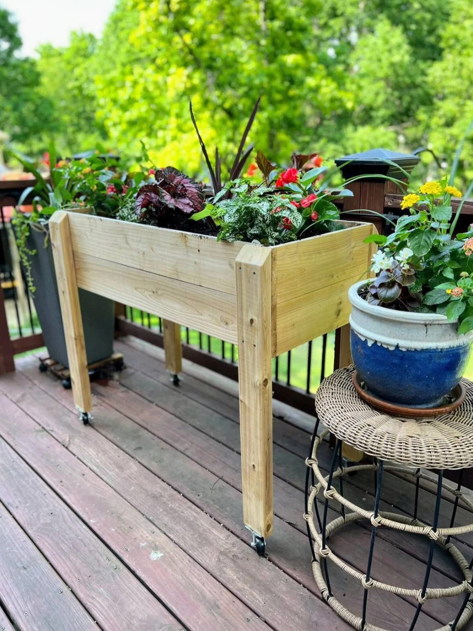 A wooden raised planter box with assorted plants is on a wooden deck. Nearby are a gray pot with greenery and a blue ceramic pot with flowers on a wicker stool. Trees in background.