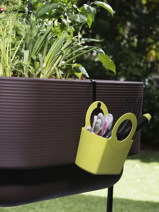 Raised garden bed with various vegetables and herbs growing, and a yellow container holding gardening tools hanging on the side.