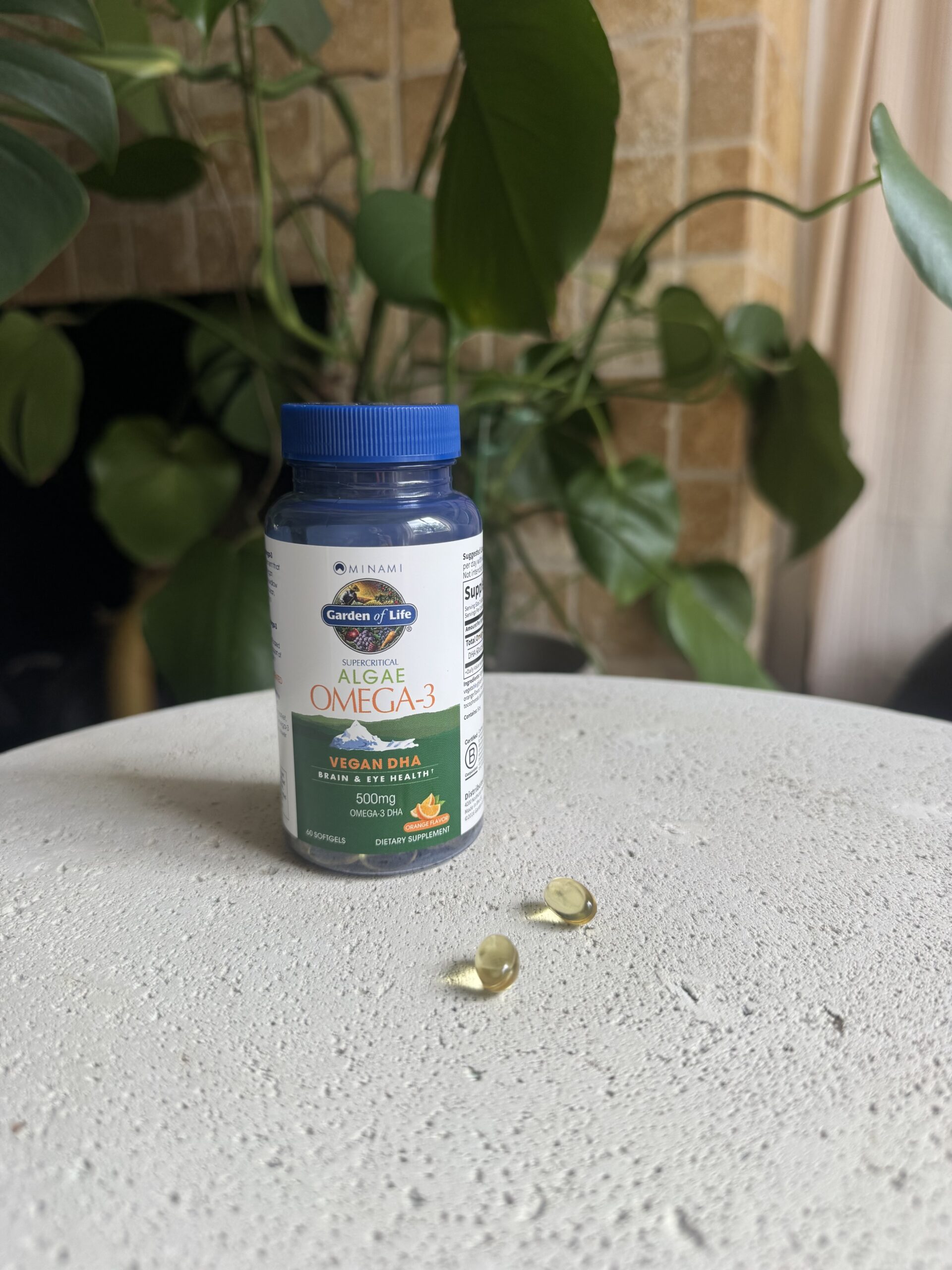 A bottle of Garden of Life Algae Omega-3 supplements sits on a round white surface with two softgel capsules beside it. Green leafy plants and a brick wall are in the background.