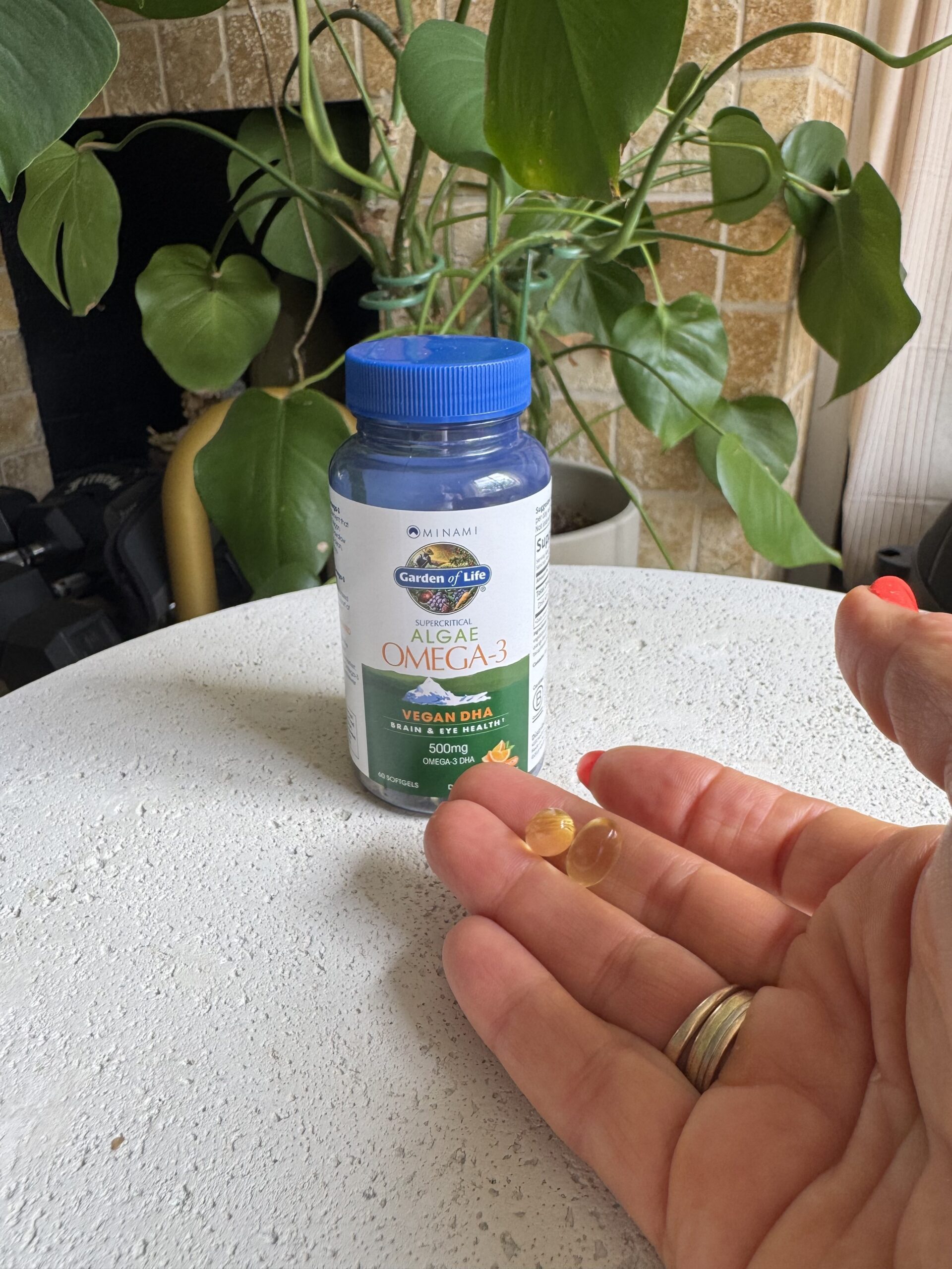 A hand holds two softgel capsules in front of a bottle labeled "Garden of Life Algae Omega-3" on a white table with a green plant in the background.
