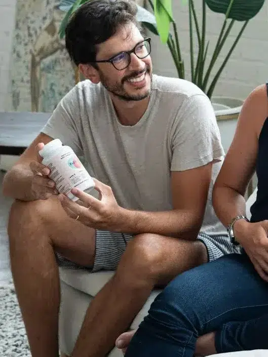 A pregnant woman sits on a couch smiling, with a man holding a supplement bottle beside her and another person facing them in the foreground.