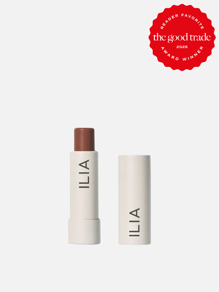 A brown ILIA lip balm with the cap removed, showing the stick, alongside its closed cap. A red badge indicates "The Good Trade 2025 Reader Favorite Award Winner.