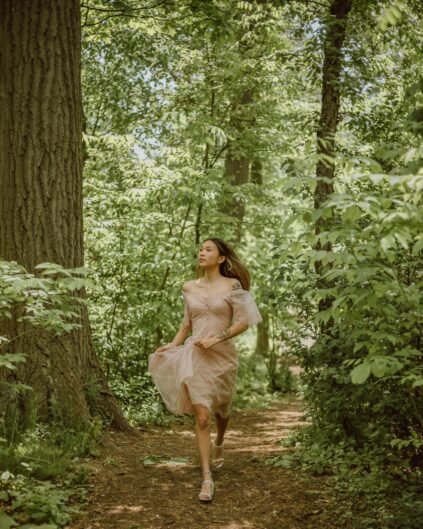 A woman in a light pink dress walks along a narrow, wooded path surrounded by dense green foliage and tall trees.
