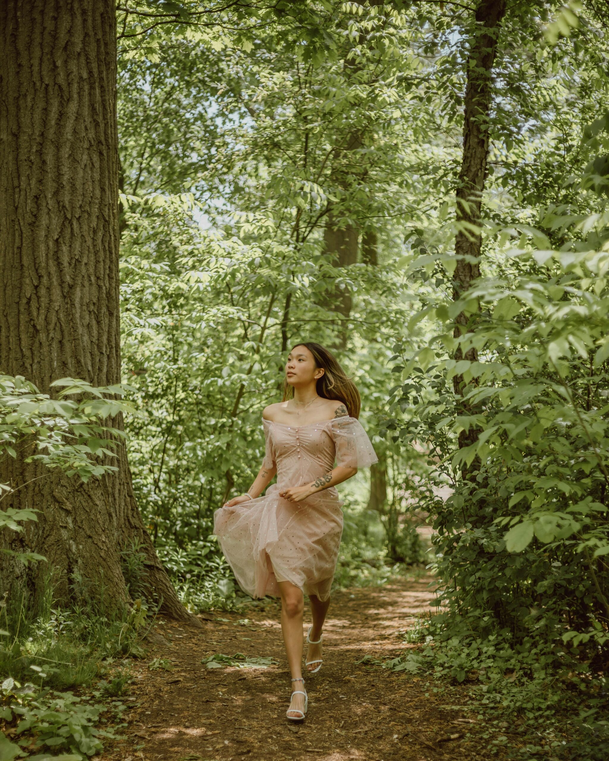 A woman in a light pink dress walks along a narrow, wooded path surrounded by dense green foliage and tall trees.