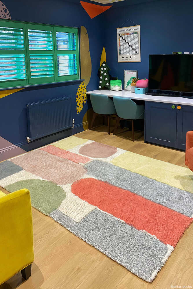 A colorful modern room with a geometric patterned rug, green window shutters, a TV on a blue cabinet, and a desk with two chairs against a dark blue wall.