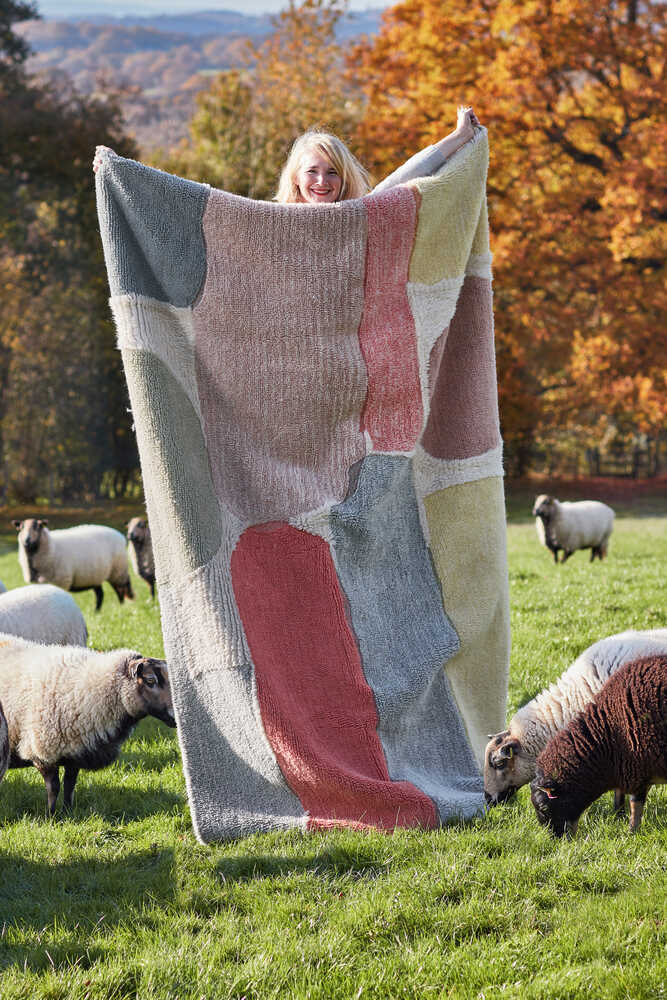 A person stands in a grassy field holding a large, multicolored blanket, surrounded by several sheep with autumn trees in the background.