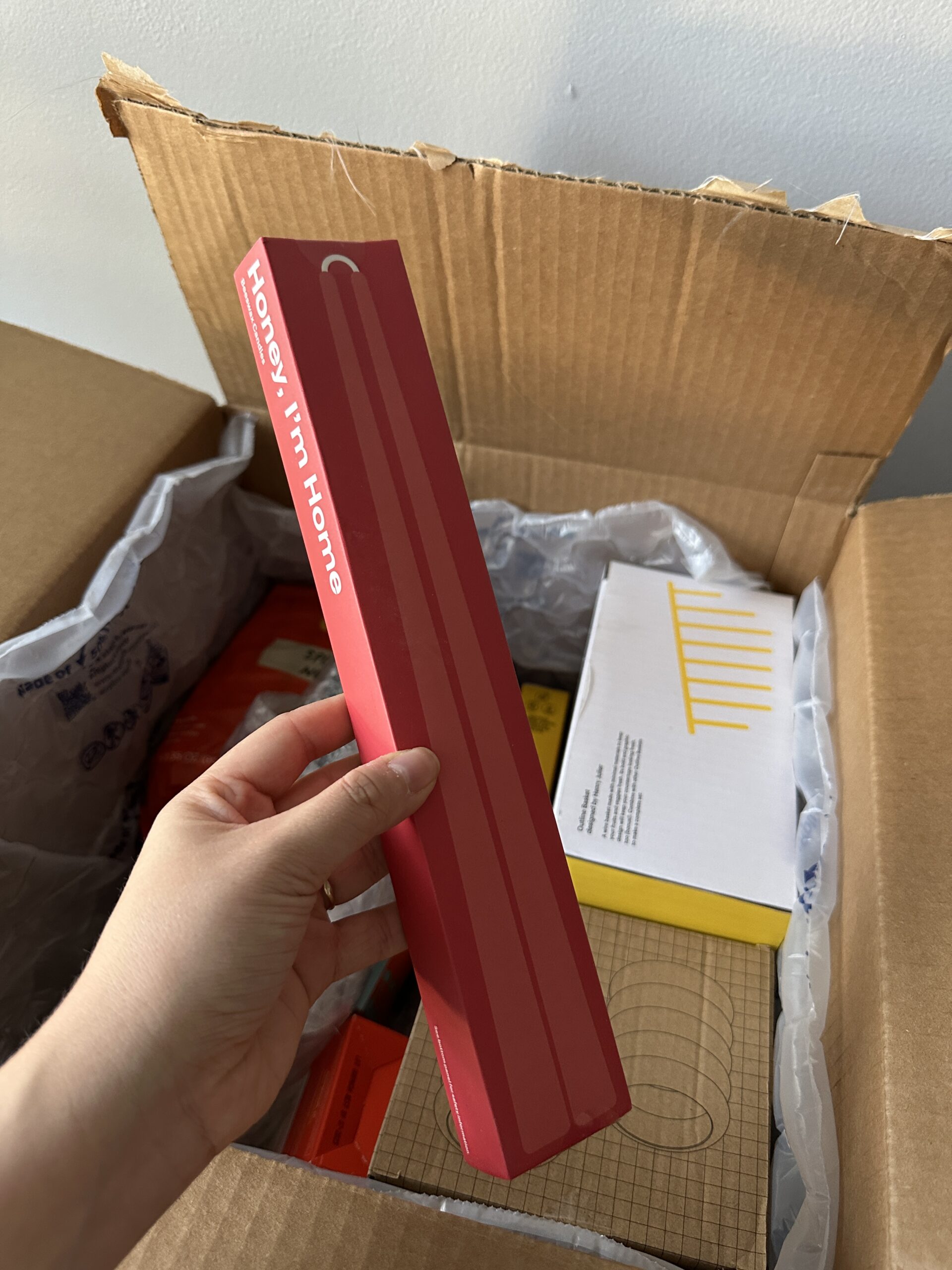 A hand holds a red rectangular box labeled "Honey, I'm Home" above an open cardboard box containing various other boxed items.