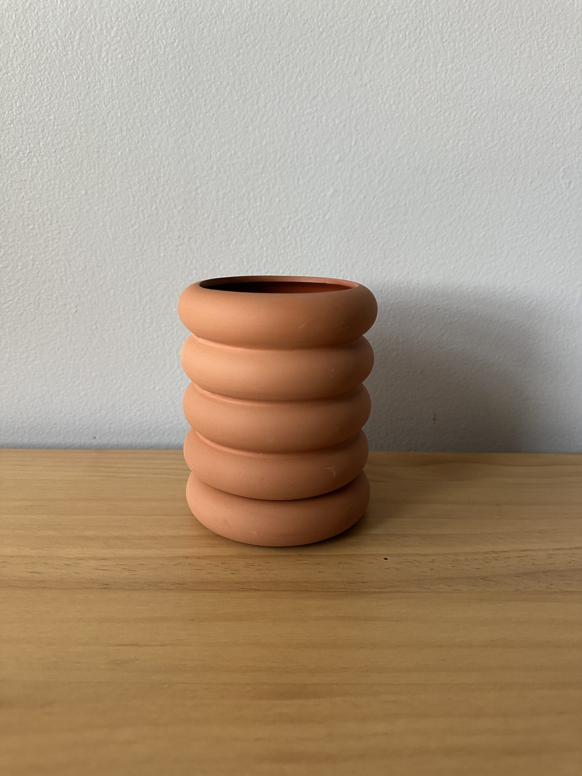 A small, round, terracotta vase with a stacked ring design sits on a wooden surface against a plain light-colored wall.