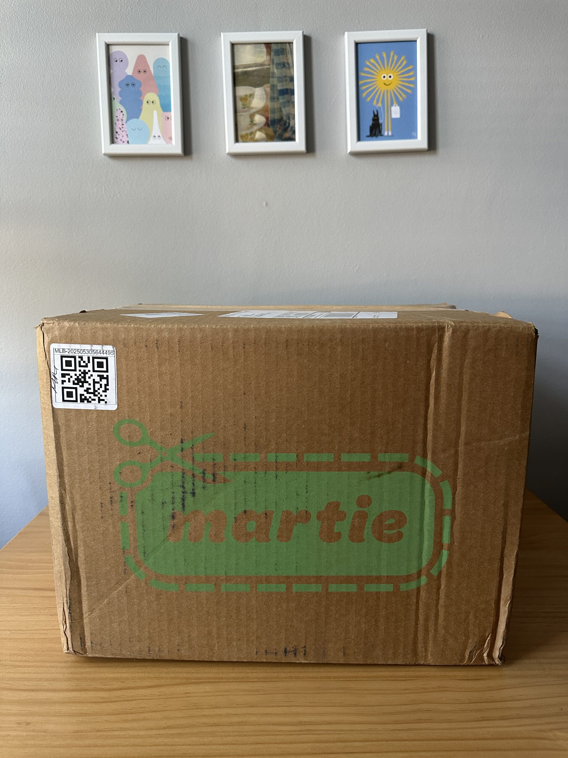 A cardboard box labeled "martie" sits on a wooden surface. Three framed pictures are hanging on a gray wall in the background.