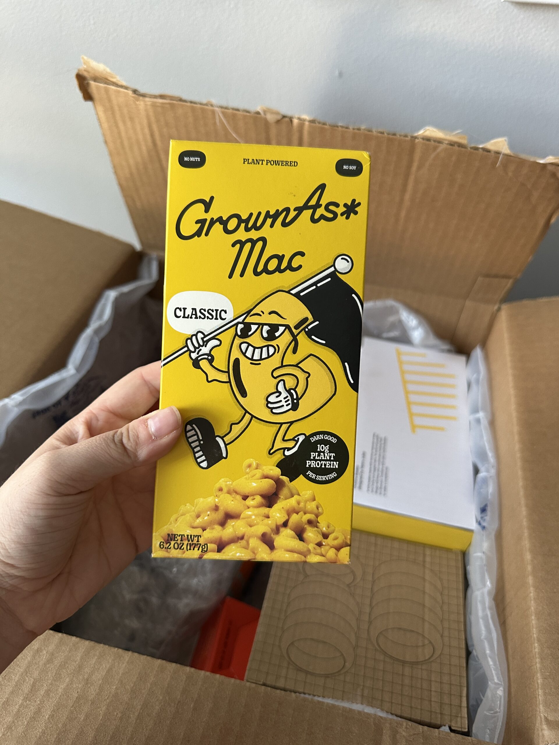 A hand holds a box of GrownAs* Mac Classic, a plant-based macaroni and cheese product, in front of an open cardboard shipping box.