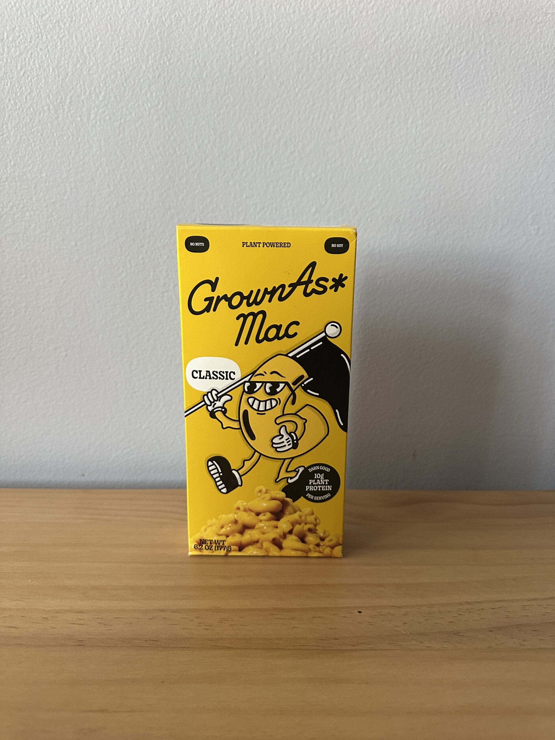 A yellow box of GrownAs* Mac Classic plant-based macaroni and cheese sits on a wooden surface against a plain light gray wall.