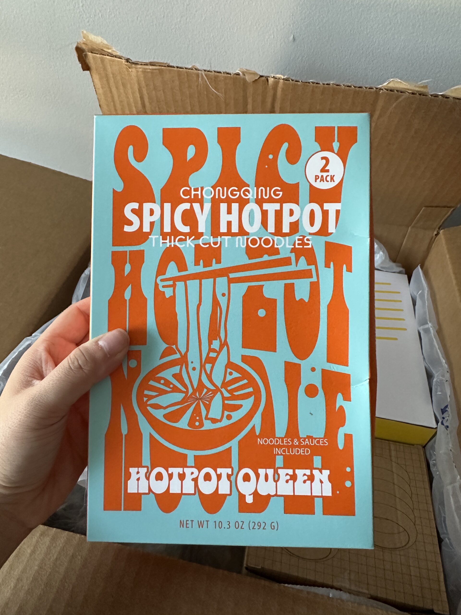 A hand holds a box labeled "Hotpot Queen Spicy Hotpot Thick Cut Noodles" with bold orange text on a blue background, next to an open cardboard box.