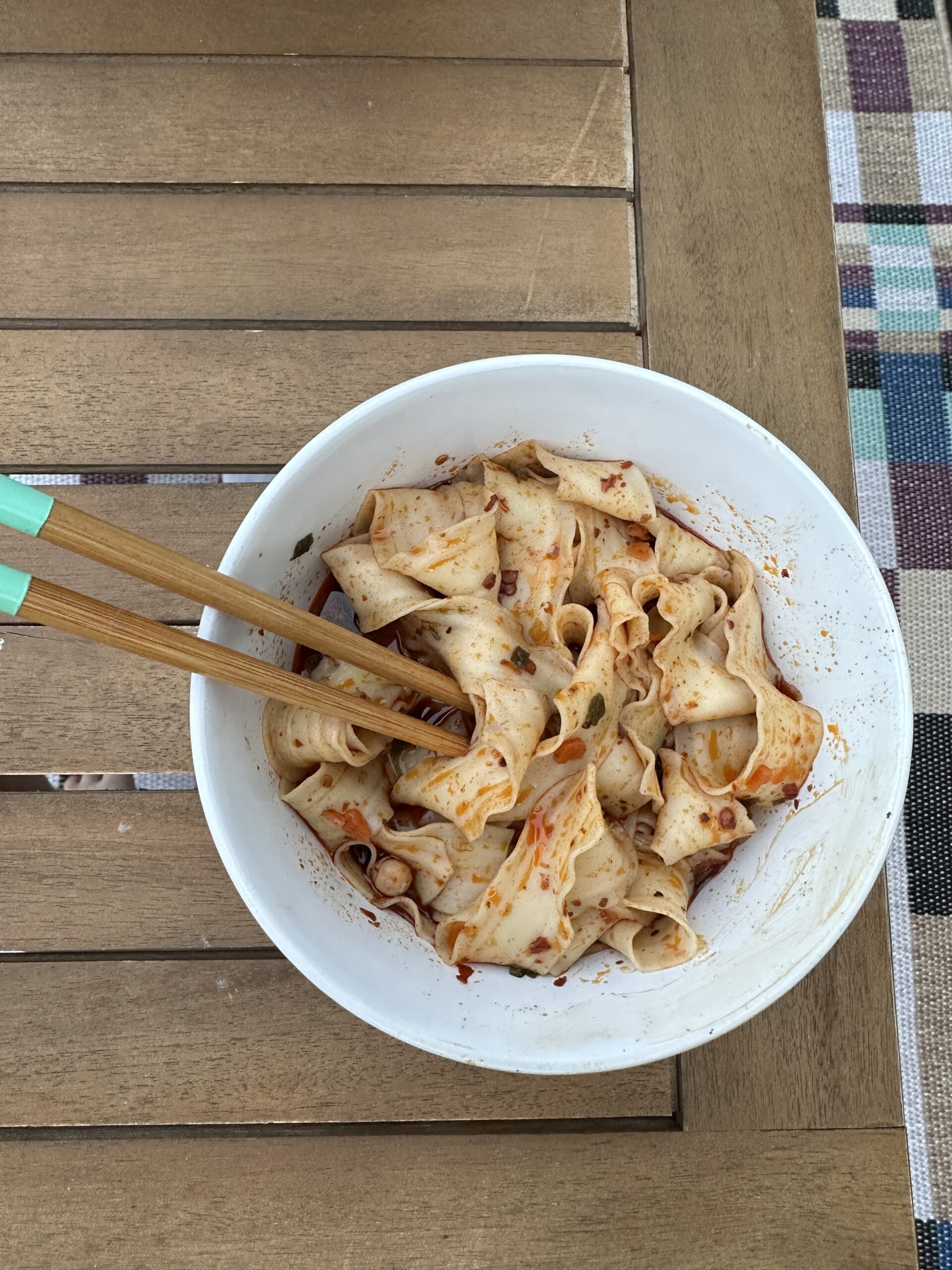 A white bowl of wide, flat noodles with chili oil and spices, accompanied by wooden chopsticks with turquoise grips, sits on a wooden table.