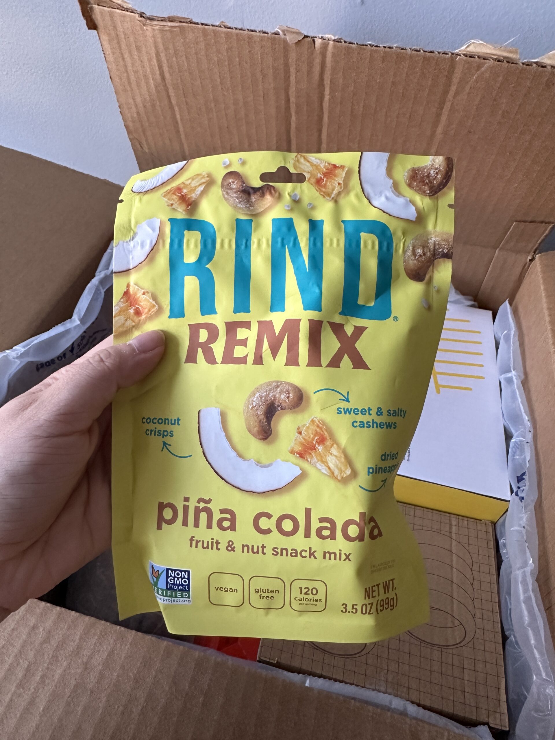 A hand holds a yellow bag of RIND REMIX piña colada fruit and nut snack mix in front of an open cardboard box.