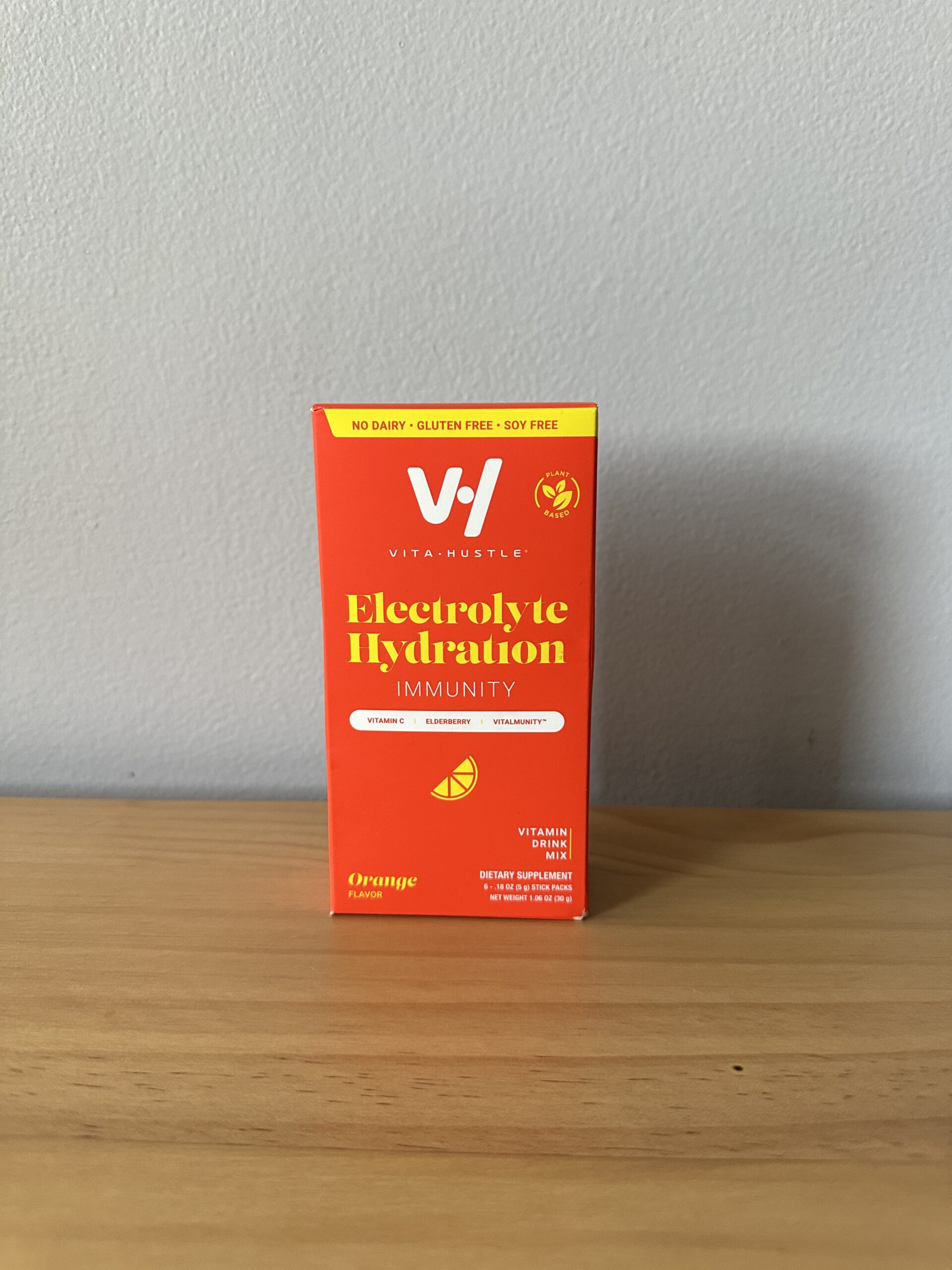 A box of Vita-Muscle Electrolyte Hydration Immunity supplement in orange flavor is placed on a wooden surface against a plain light gray wall.
