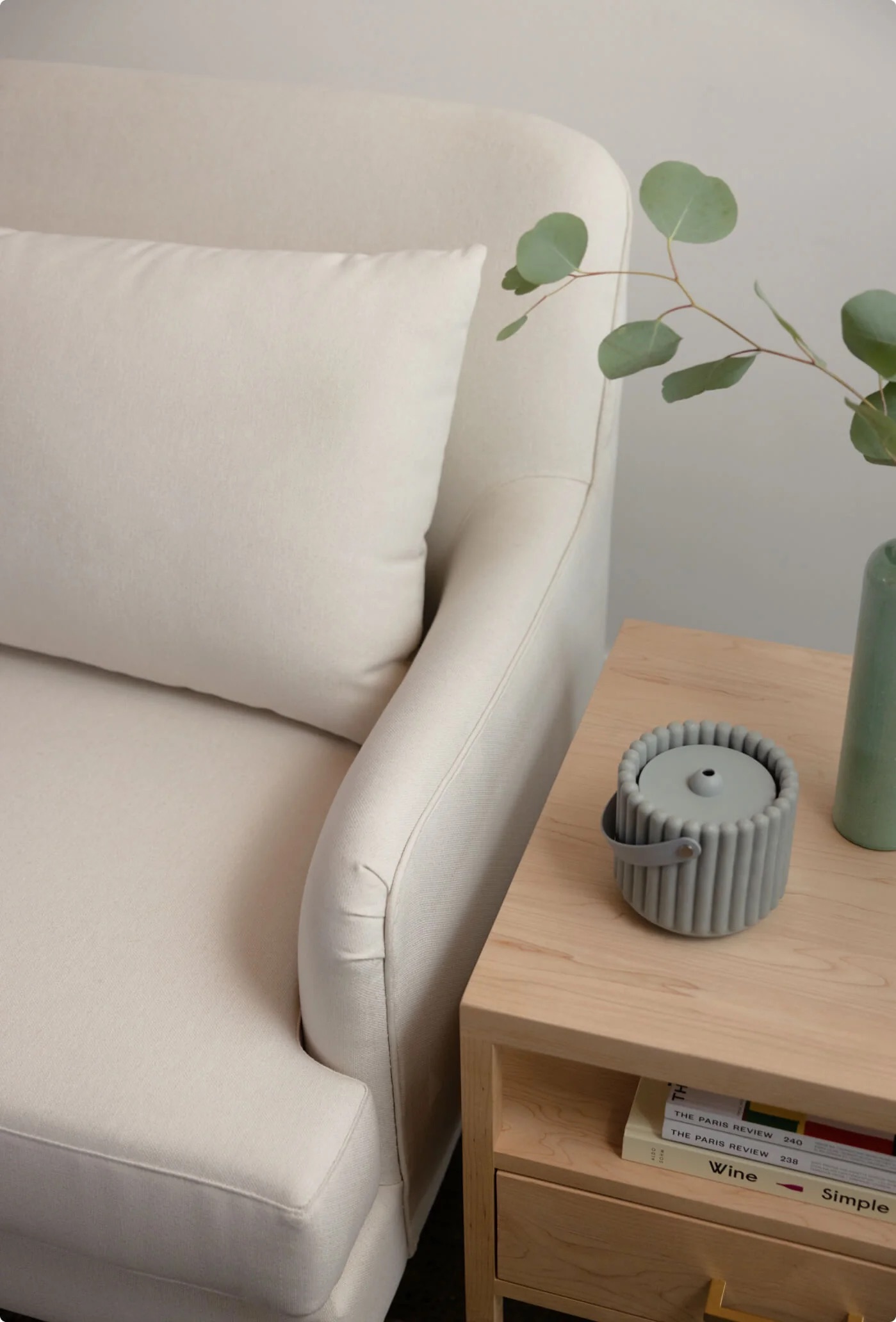 A beige sofa next to a light wood side table holding a green vase with eucalyptus branches and a gray candle in a ribbed holder.