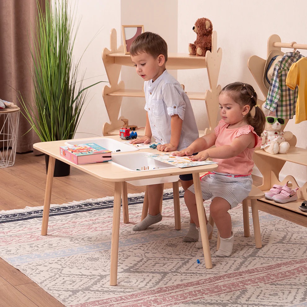 Two young children sit at a wooden table in a playroom, drawing and playing with toys. Shelves, clothes, and stuffed animals are visible in the background.