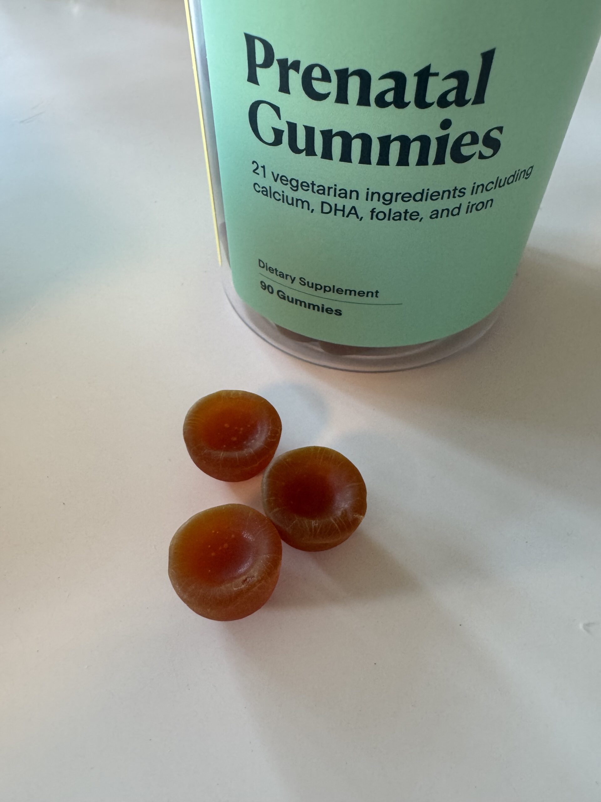 Three round, brown gummies are placed on a white surface next to a container labeled "Prenatal Gummies," highlighting these as a convenient and tasty prenatal vitamin option for expecting mothers.