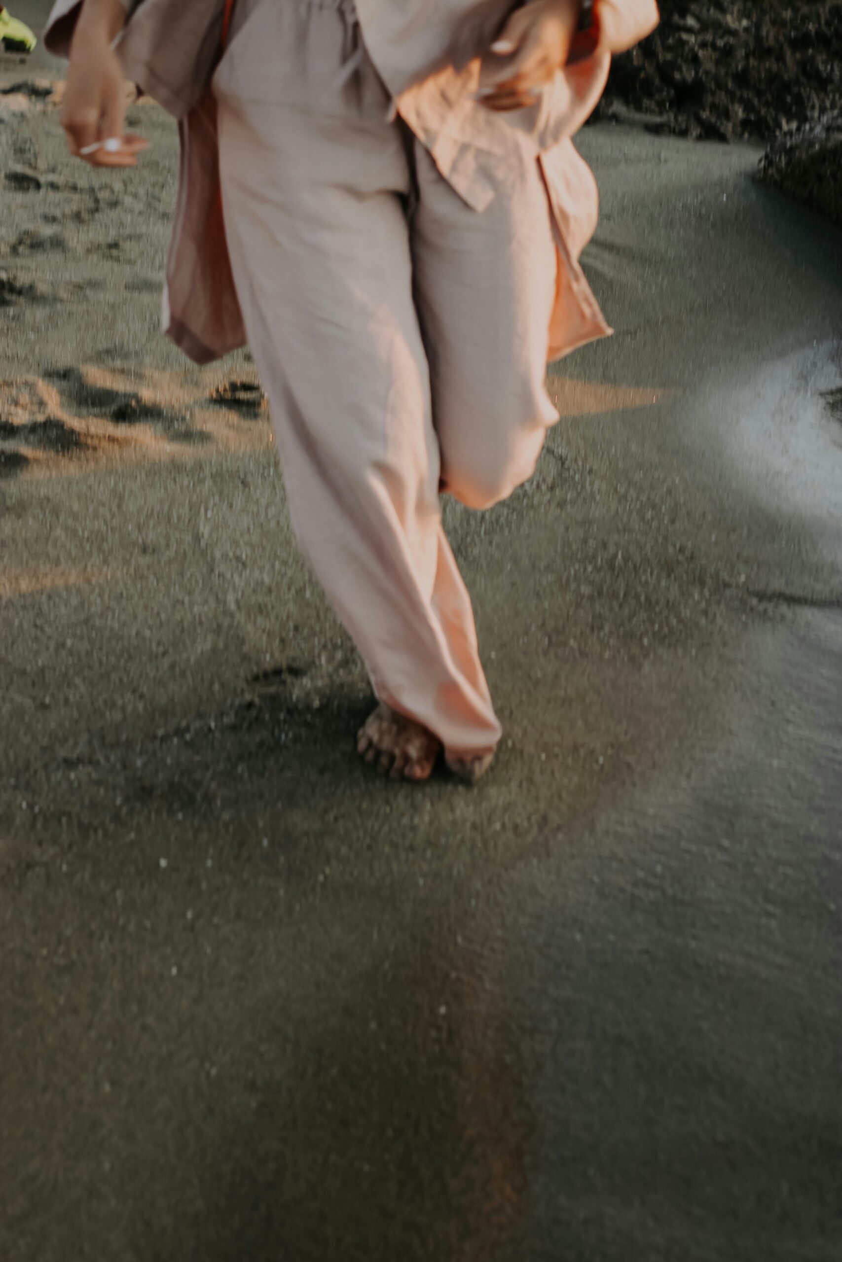 A person wearing loose, light pink clothing walks barefoot along the wet sand at the beach.