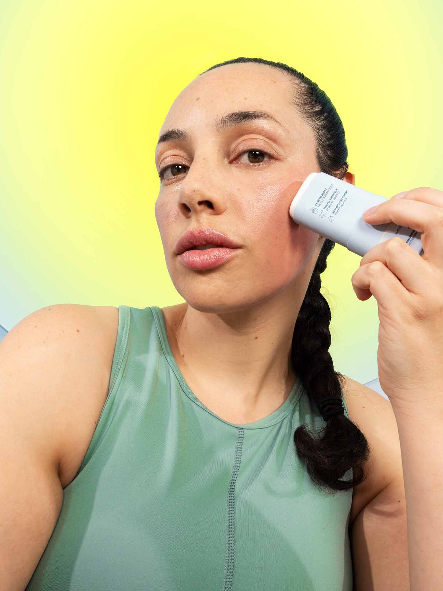 A person in a green tank top applies a stick cosmetic product to their cheek, with a yellow sun-like backdrop behind them.