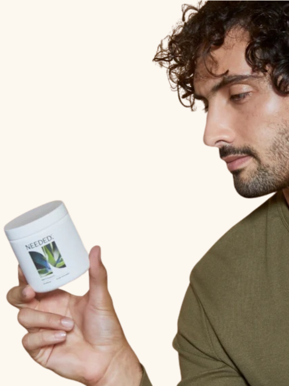 A man with curly hair and a green shirt looks closely at a white jar labeled "NEEDO" with a plant image on it.