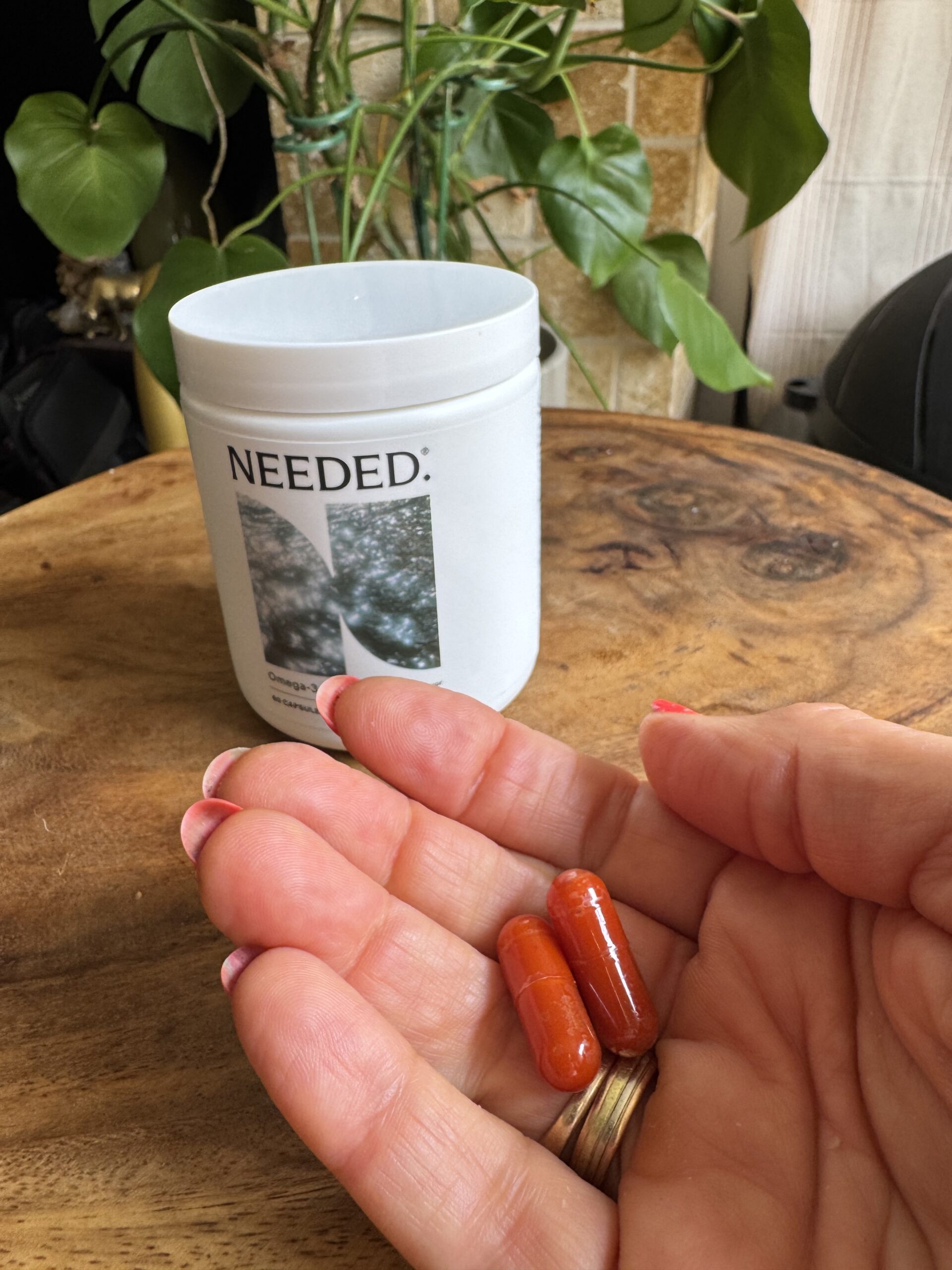 A hand holding two red capsules in front of a white container labeled "NEEDED:" on a wooden table, with a green plant in the background.