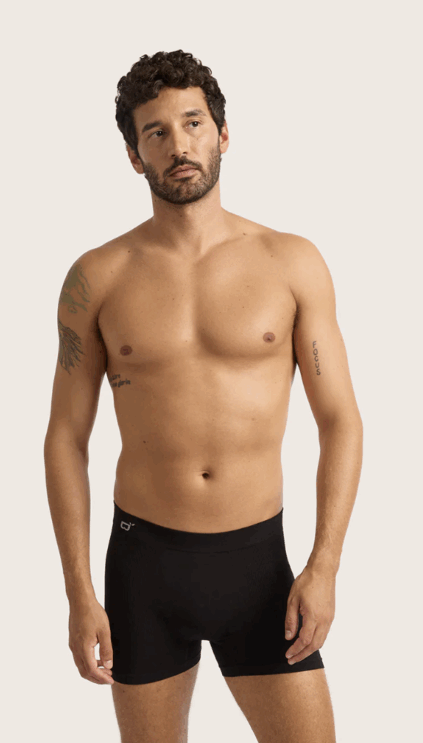 A shirtless man with tattoos is standing and wearing black boxer briefs against a plain light background.