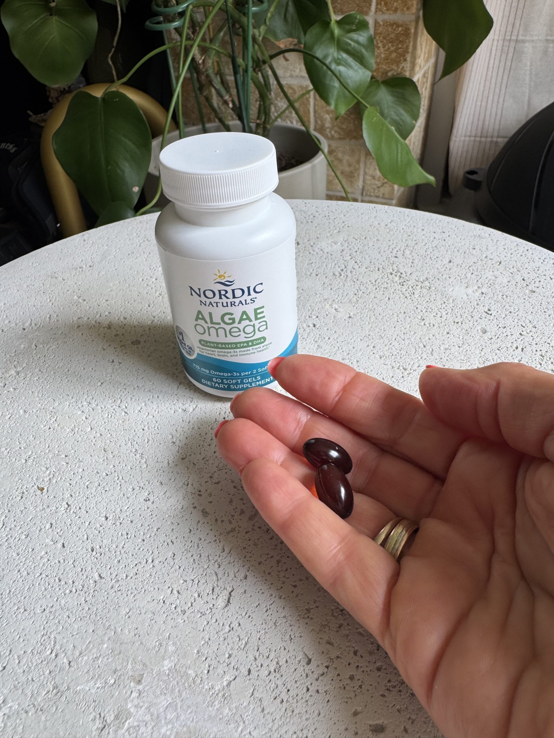 A hand holds three dark softgel capsules in front of a bottle labeled "Nordic Naturals Algae Omega" on a white table with green plants in the background.
