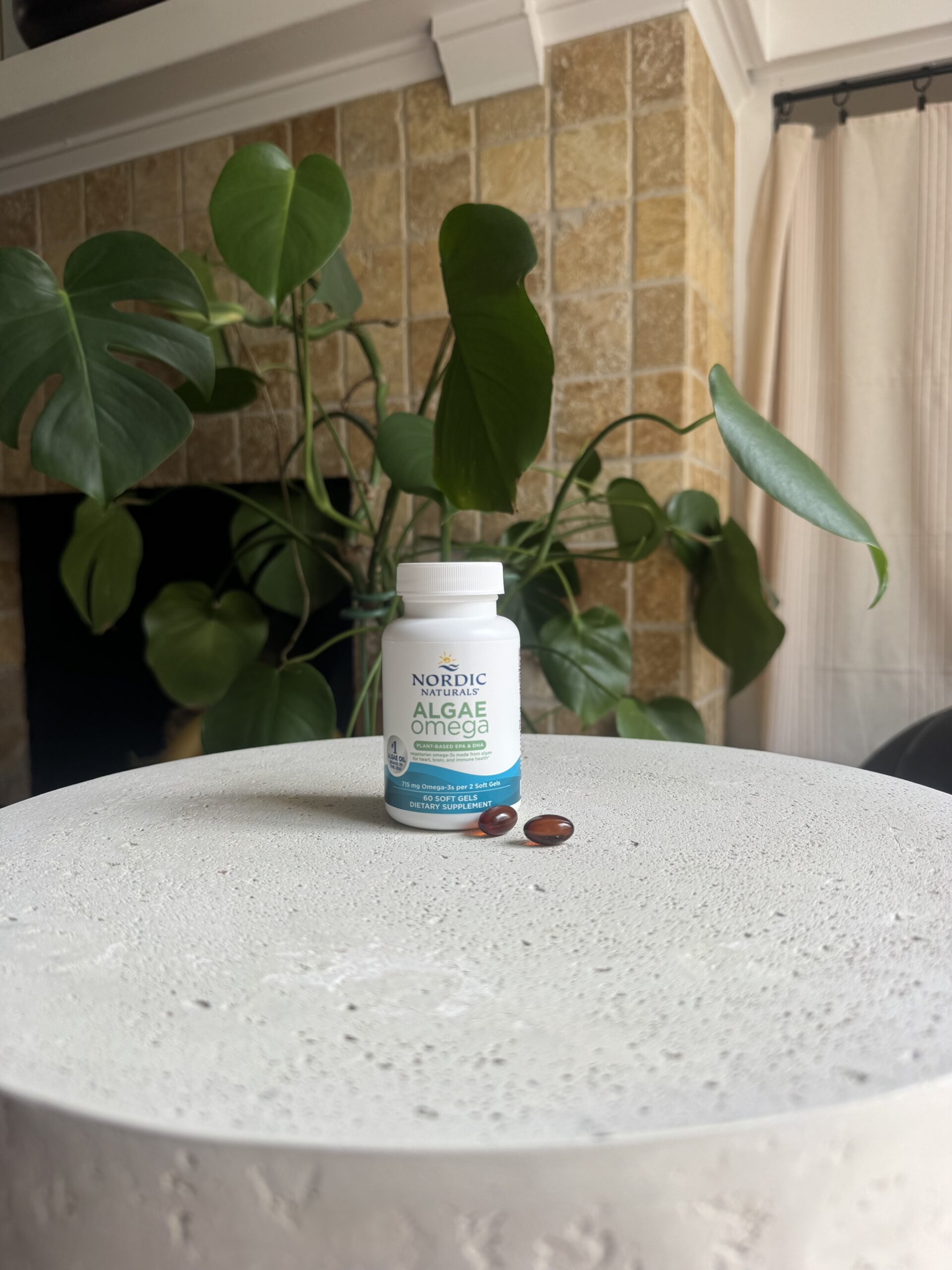 A bottle of Nordic Naturals Algae Omega supplement with two softgels on a round gray table, set in front of green plants and a tiled fireplace.
