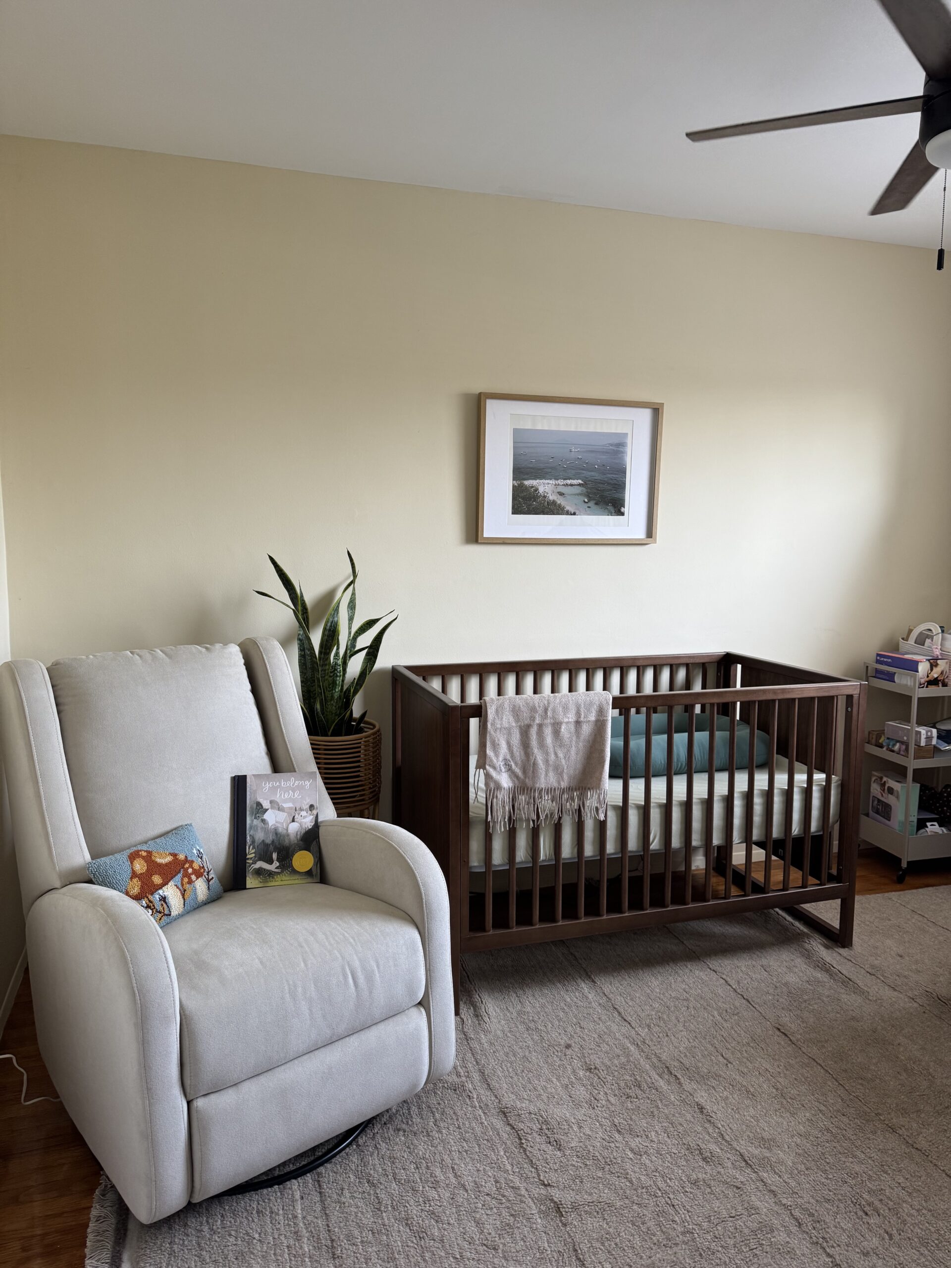 A nursery with a crib, a white recliner, a potted plant, a framed photo on the wall, and a small shelf with baby items.