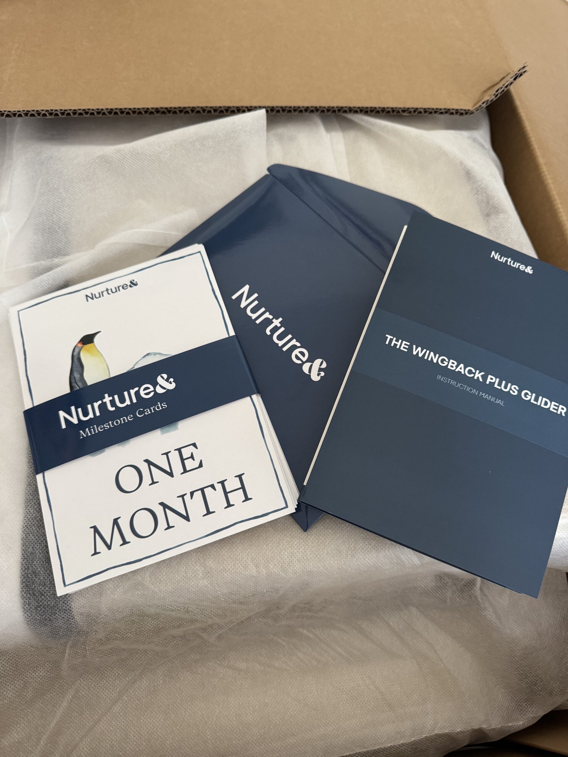 A box contains Nurture& milestone cards, an instruction manual for "The Wingback Plus Glider," and a blue folder, all placed on top of packing material.