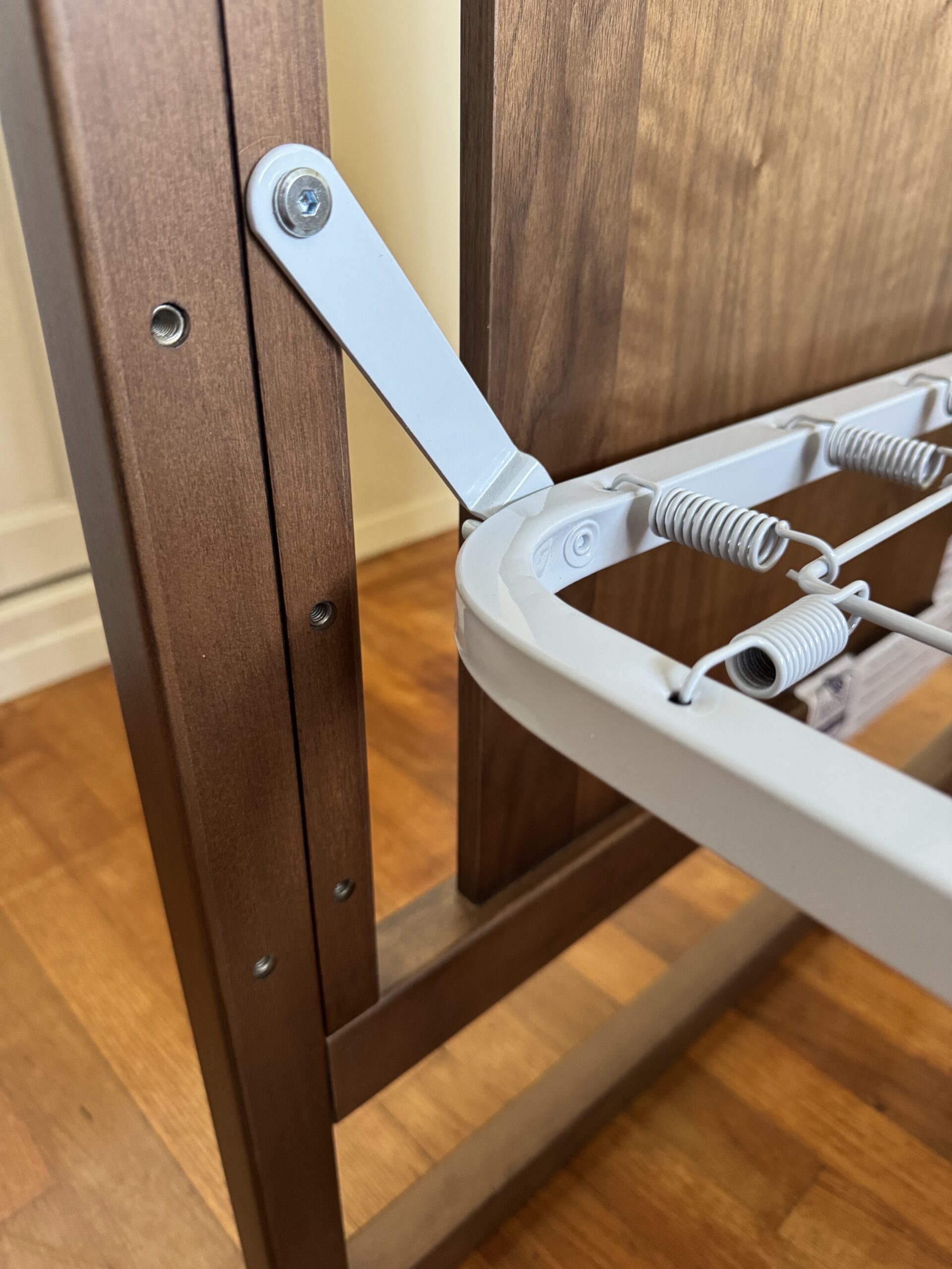 Close-up of a metal folding mechanism attached to a wooden frame, likely part of a piece of convertible furniture, on a hardwood floor.