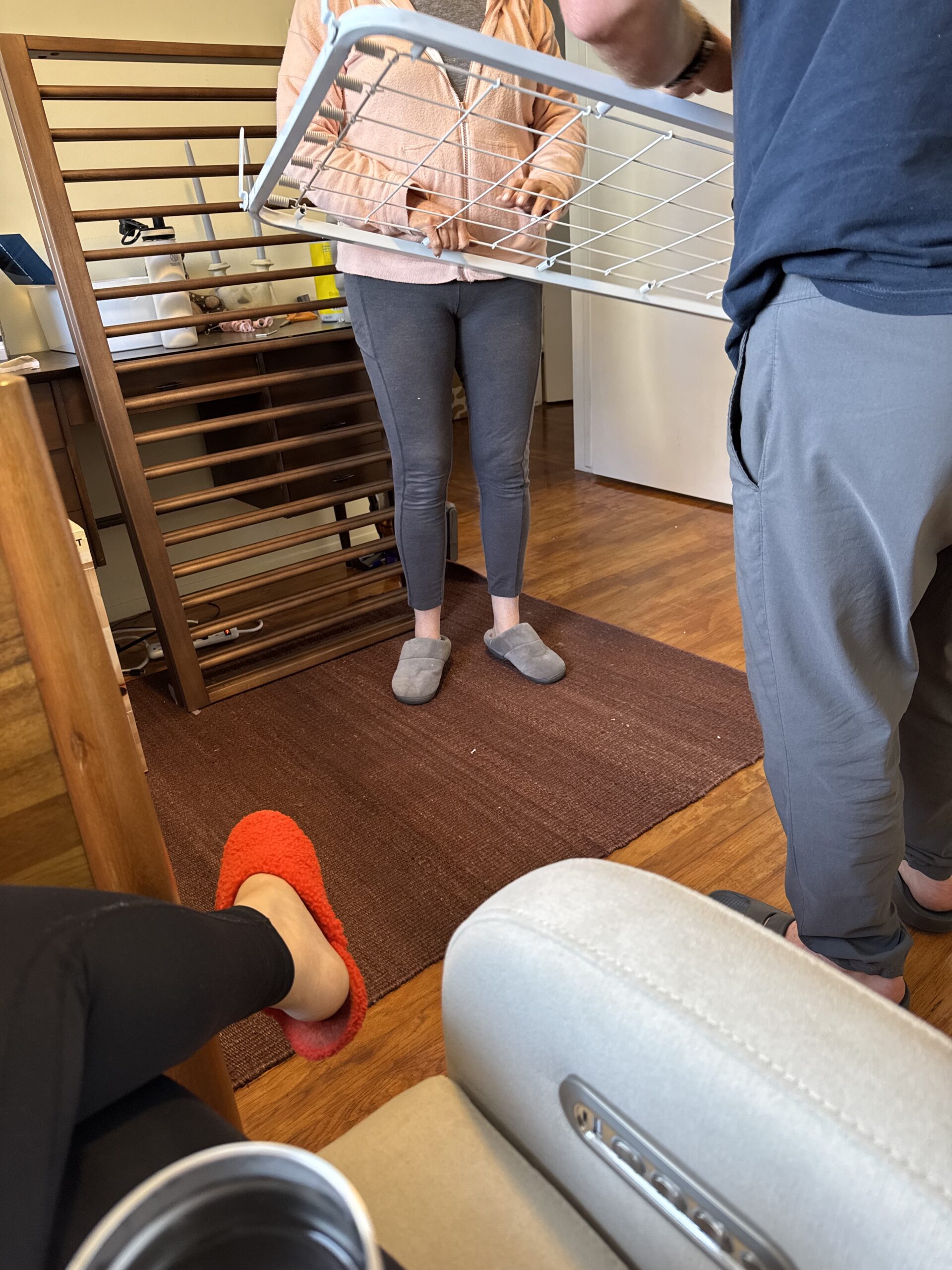 Two people stand assembling furniture in a room with wooden floors, while another person sits nearby wearing red slippers. A drying rack and furniture parts are visible.