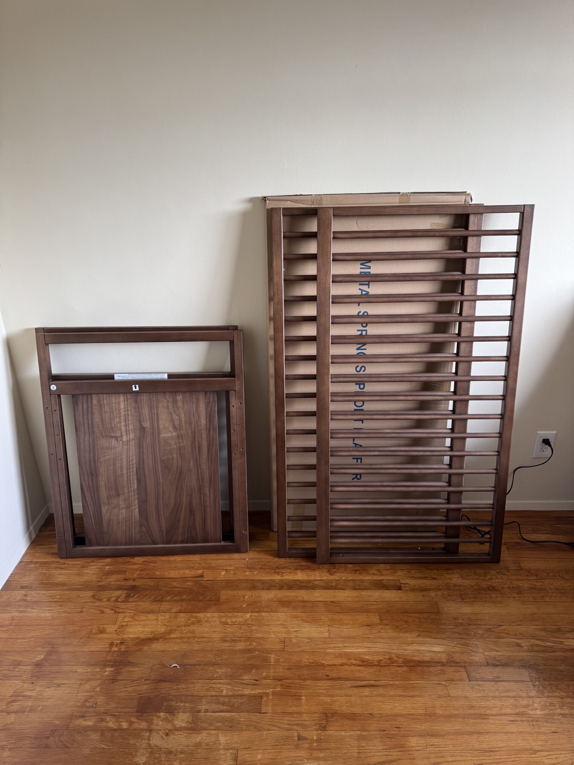 Two wooden pieces of furniture, likely parts of a crib or bed frame, are leaning against a beige wall on a wooden floor next to an electrical outlet.