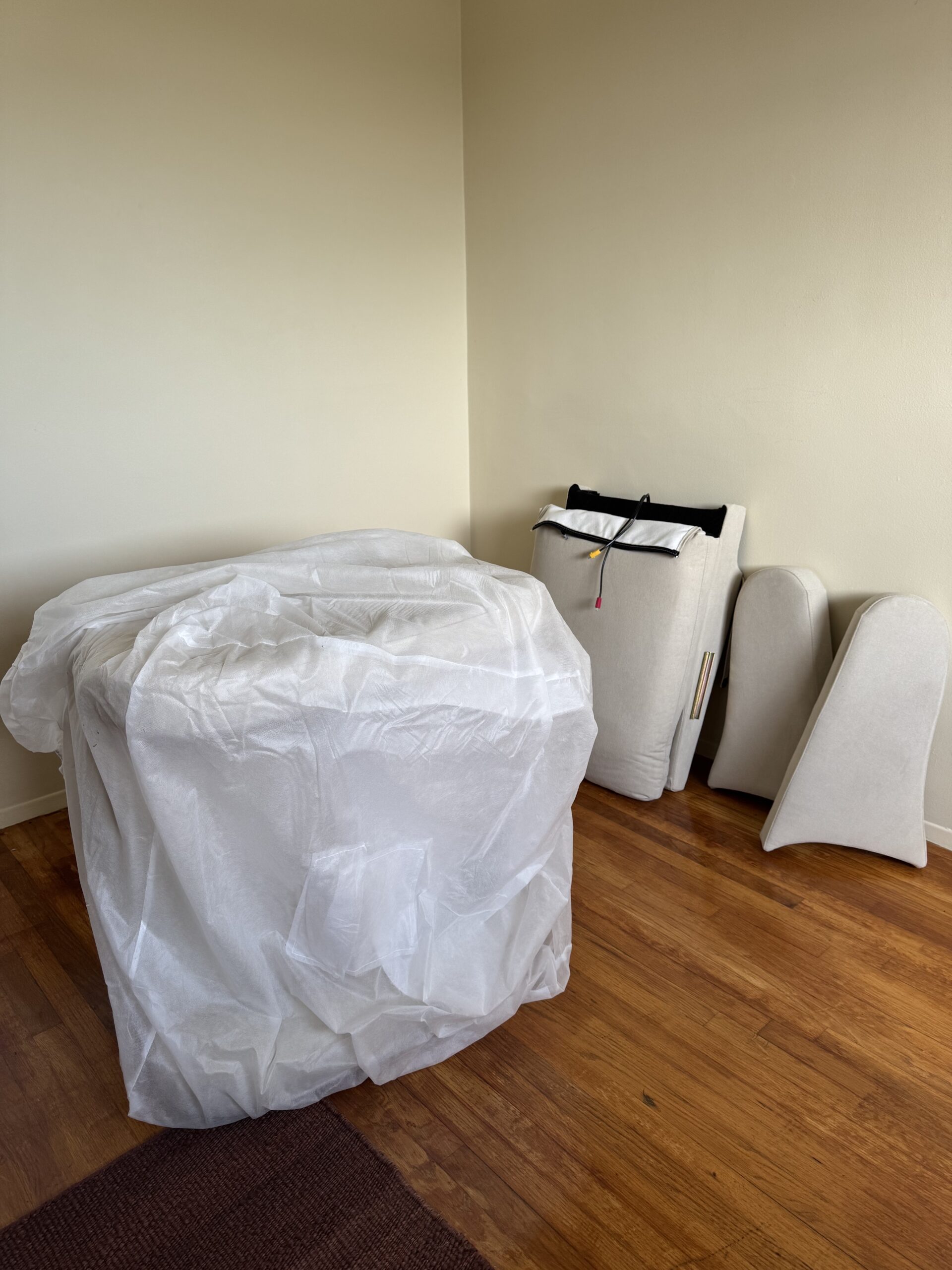 Disassembled sofa parts, including cushions and fabric-wrapped pieces, are placed on a hardwood floor in an empty room with beige walls.