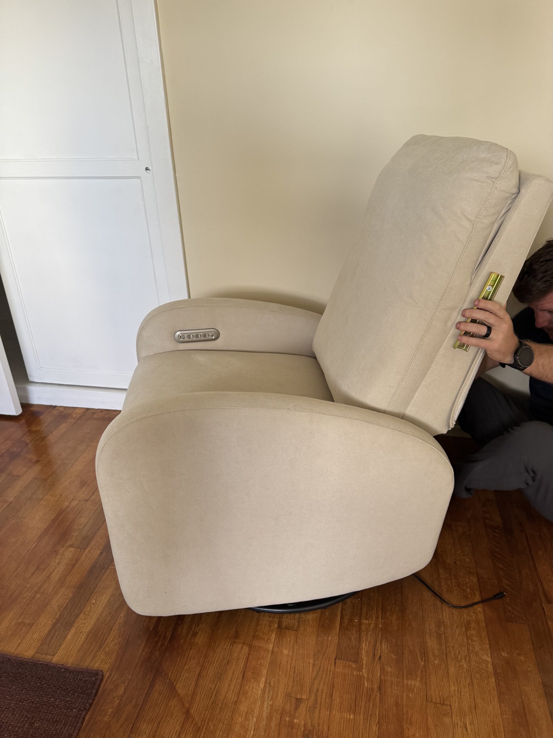 A person measuring the gap behind the backrest of a beige upholstered recliner chair on a wooden floor near a cream-colored wall and cabinet.
