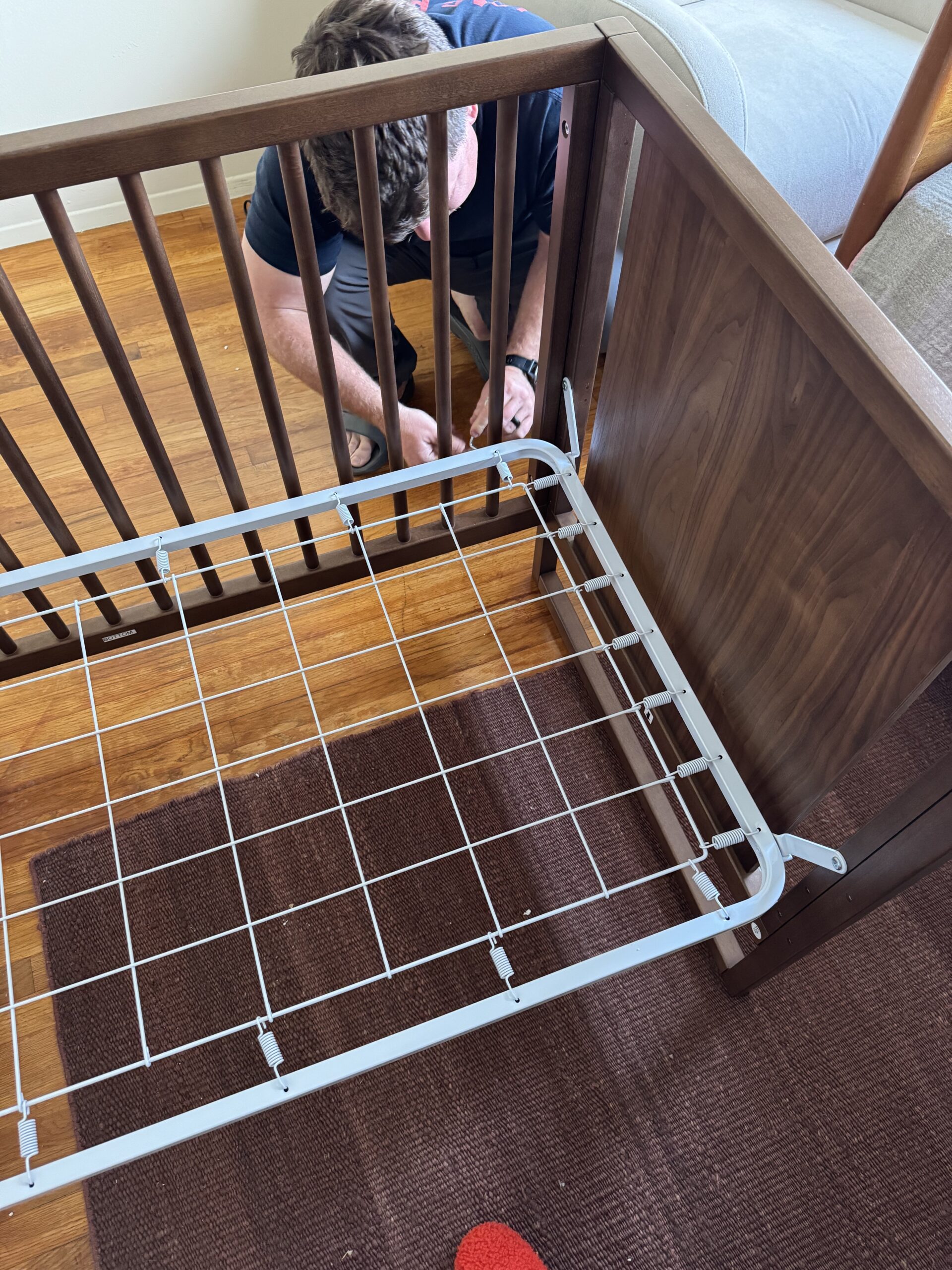 A person kneels on a wooden floor assembling or adjusting a brown wooden crib with a metal mattress support frame.