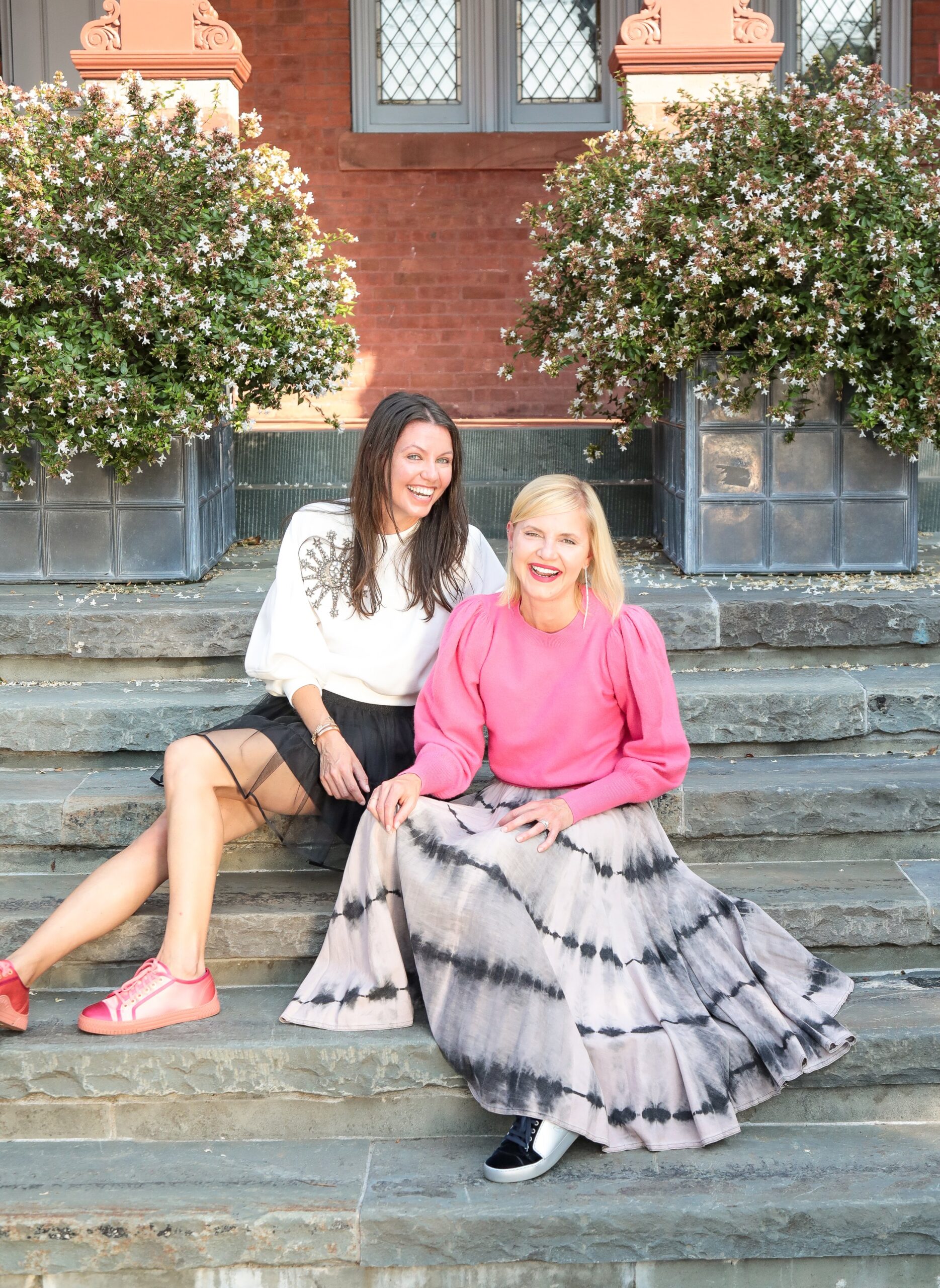 Two women sitting on outdoor stone steps, smiling. One wears a white blouse and black skirt, the other a pink sweater and tie-dye skirt. Flowering bushes and a brick building are behind them.