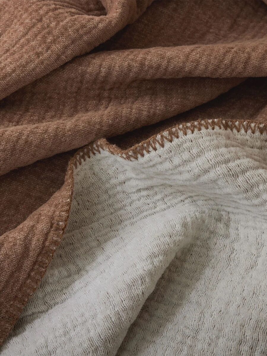 A close-up of a textured fabric blanket with brown and light beige sections, showing detailed stitching along the edge.