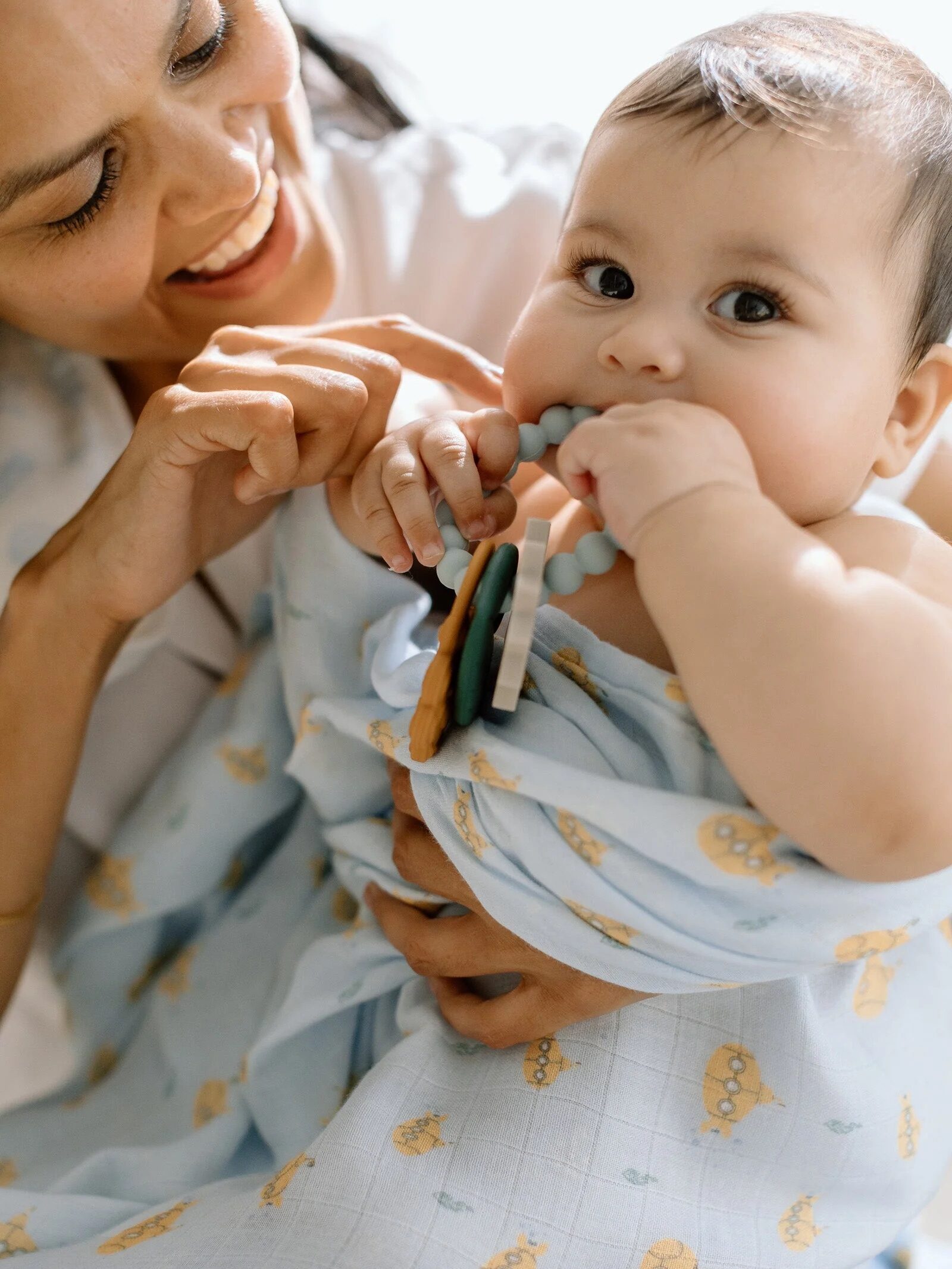 An adult smiles while holding a baby wrapped in a light blue, patterned blanket; the baby is chewing on a teething toy.