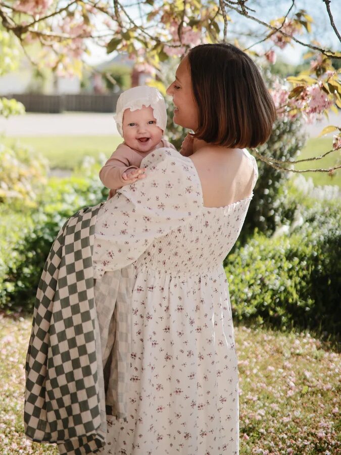A woman in a floral dress holds a smiling baby with a bonnet and checkered blanket in a sunlit garden.